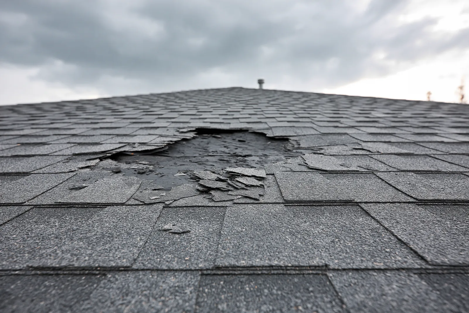 A hole in the roof shows through torn shingles under a stormy sky